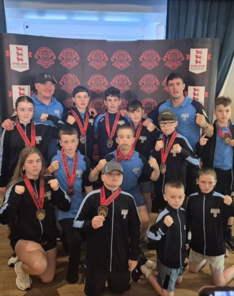 A large group of young club boxers with their coaches, wearing medals and raising fists at a box cup event.
