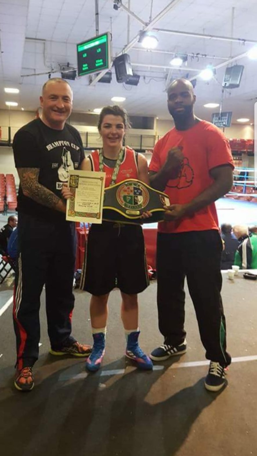 A young female boxer holding a championship belt and certificate between two coaches.